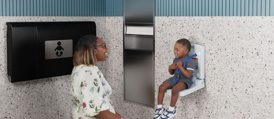 A young child sits in a toddler wall seat in a public restroom while his mother kneels nearby