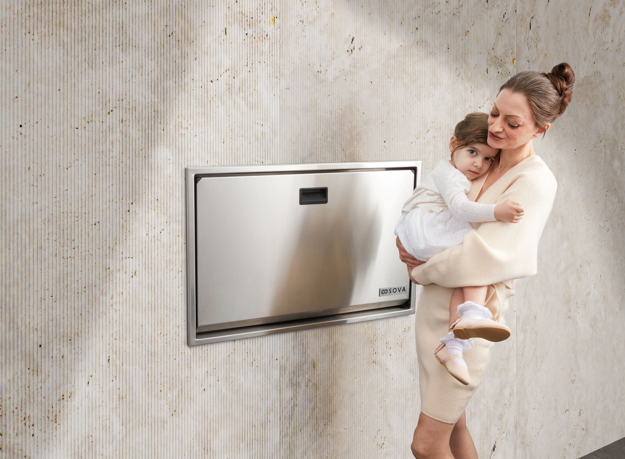 Mother holding infant next to a stainless steel baby changing table in modern restroom