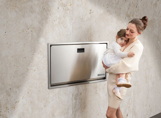 Mother holding infant next to a stainless steel baby changing table in modern restroom