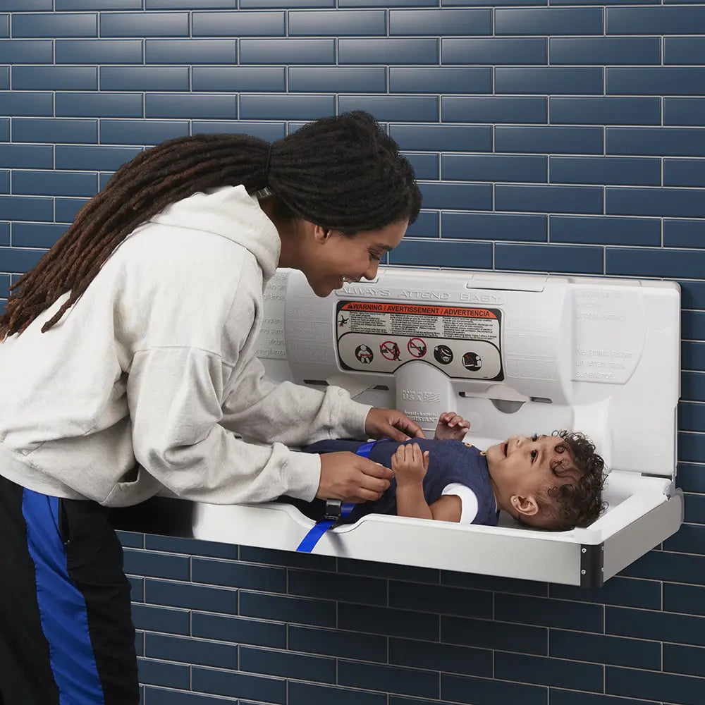Woman attending to a child in a Sova baby changing station against a blue tiled wall.