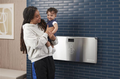 Woman holding a child next to Aura baby changing station against a blue tiled wall.