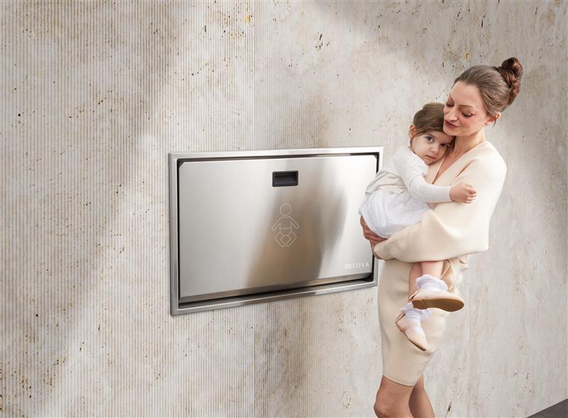 Woman holding a child next to a stainless steel summit baby changing station on a textured wall.
