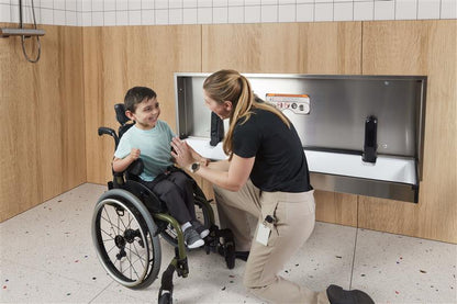 Person assisting a child in a wheelchair using Solace Extended Changing Table mounted in bathroom