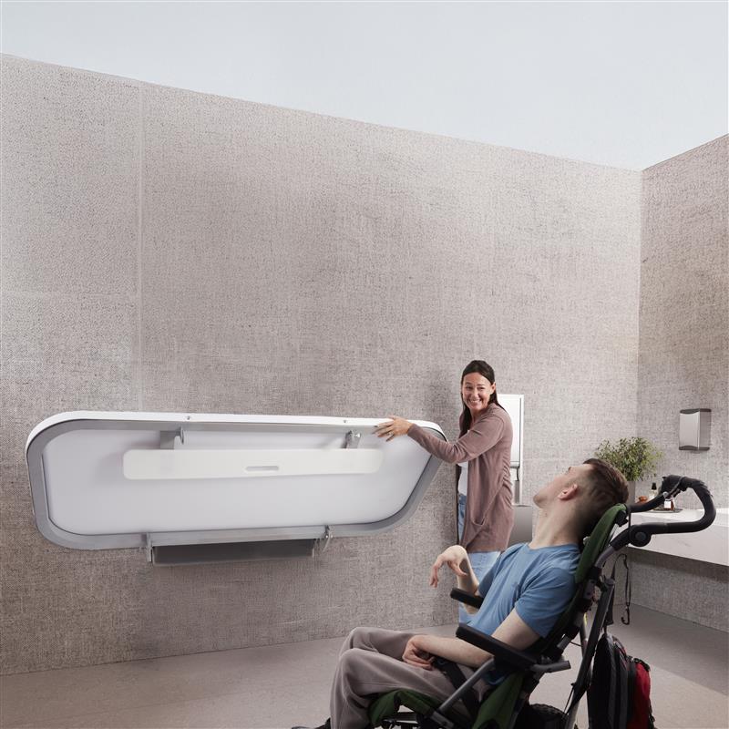 Person using a wheelchair interacting with Attend Adult Changing table in a minimalistic room.