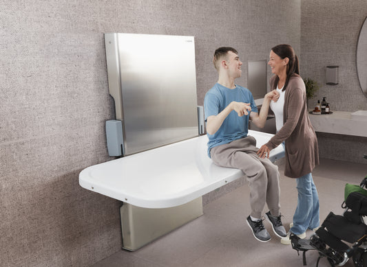 Two people sitting on a modern white adult changing table in a bathroom setting.